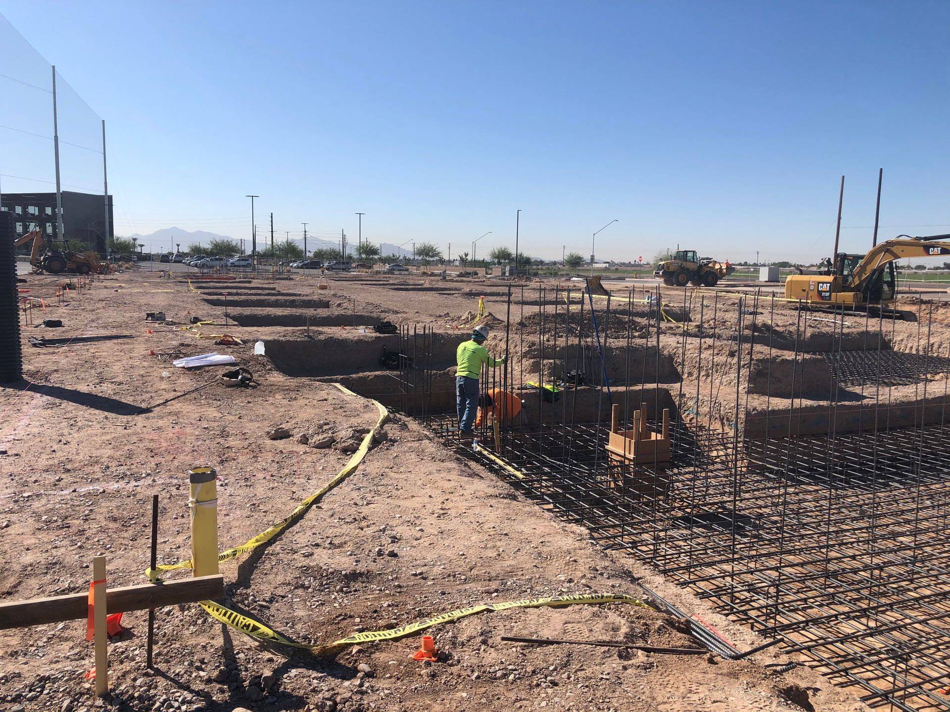 Construction site with exposed rebar, worker standing, heavy machinery, clear blue sky.