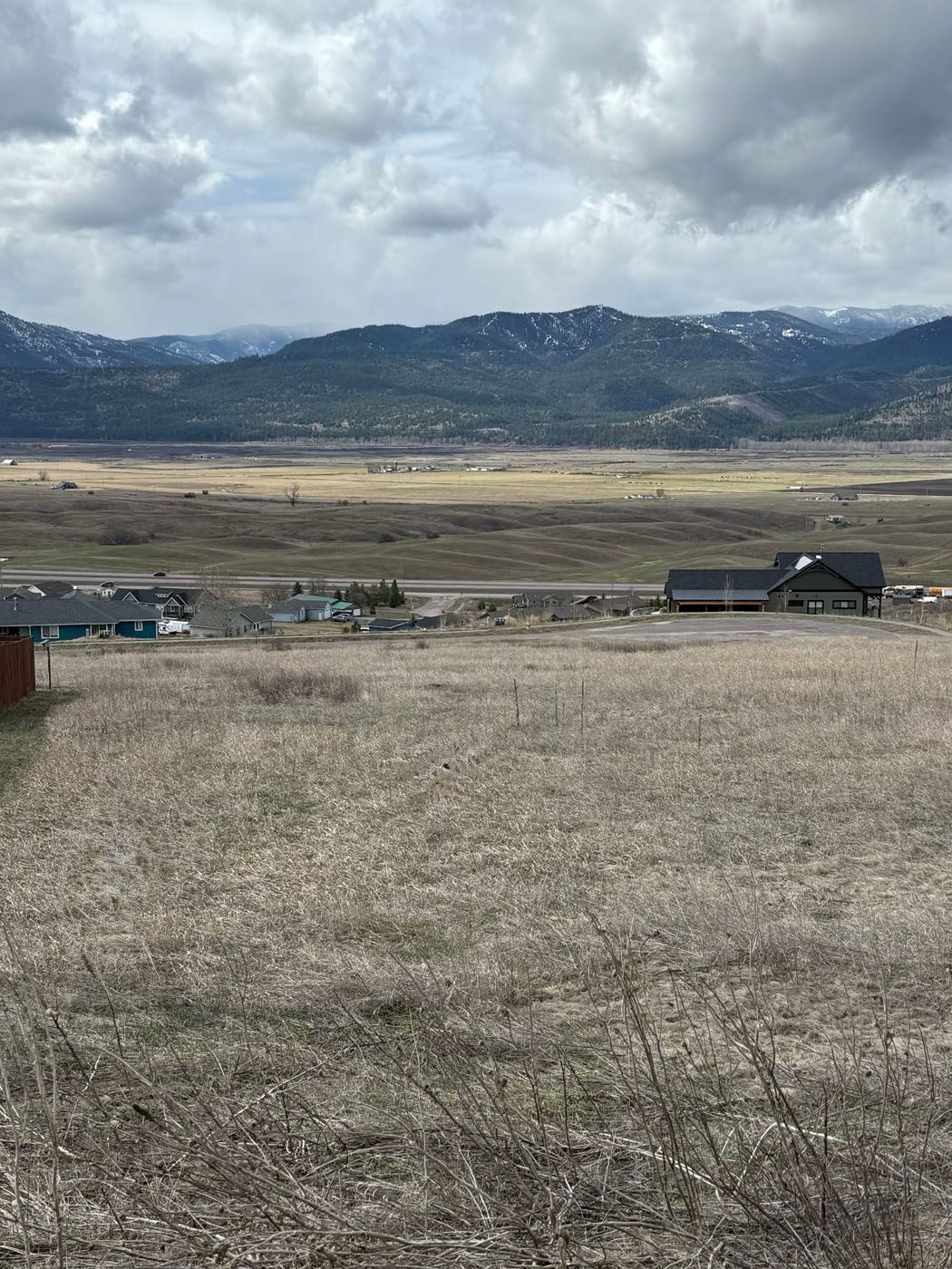 Open field view with mountains in the distance, a few buildings, and a cloudy sky.