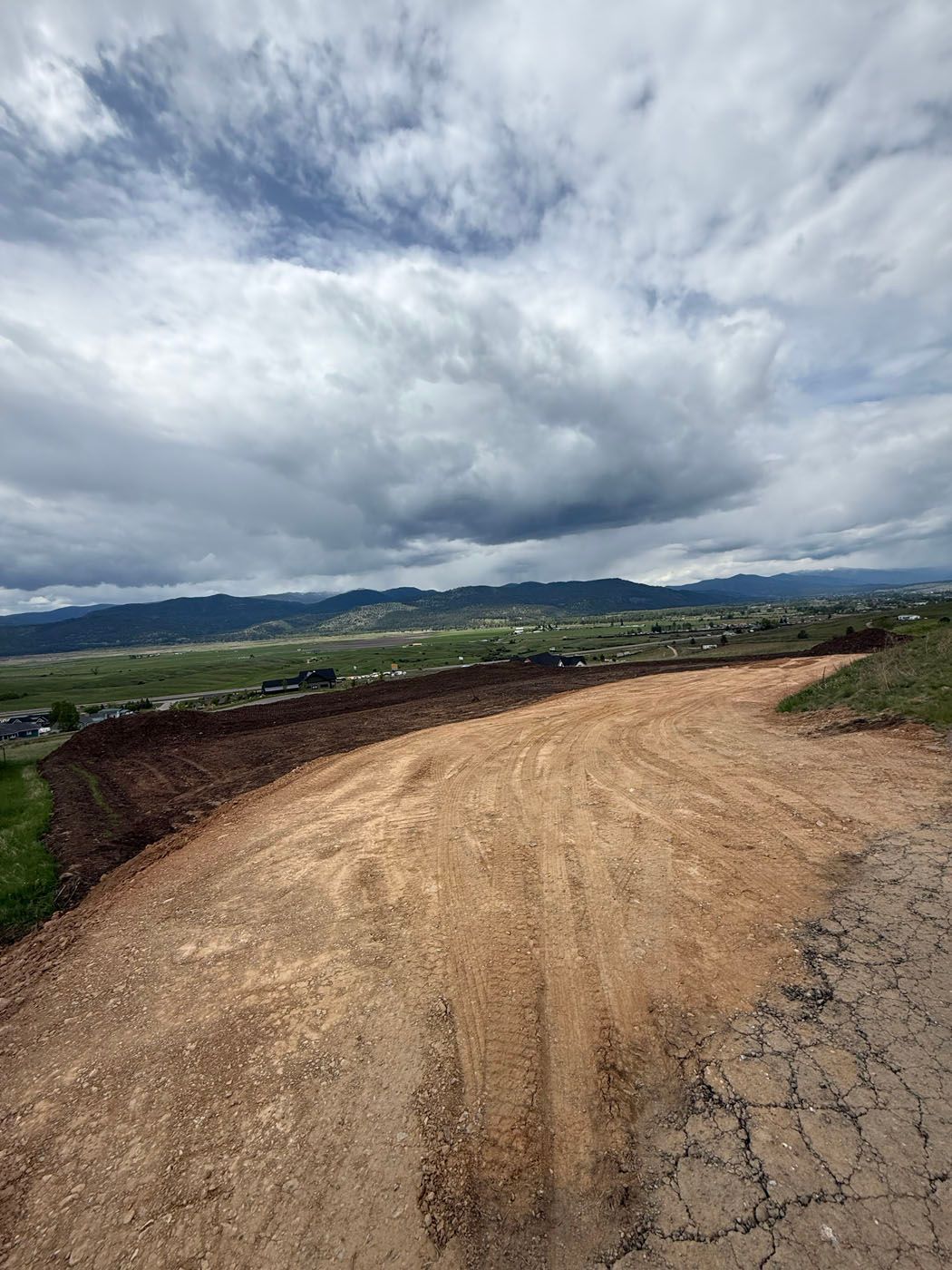 Dirt road curves uphill towards a valley under a cloudy sky. Hills and houses visible in the distance.