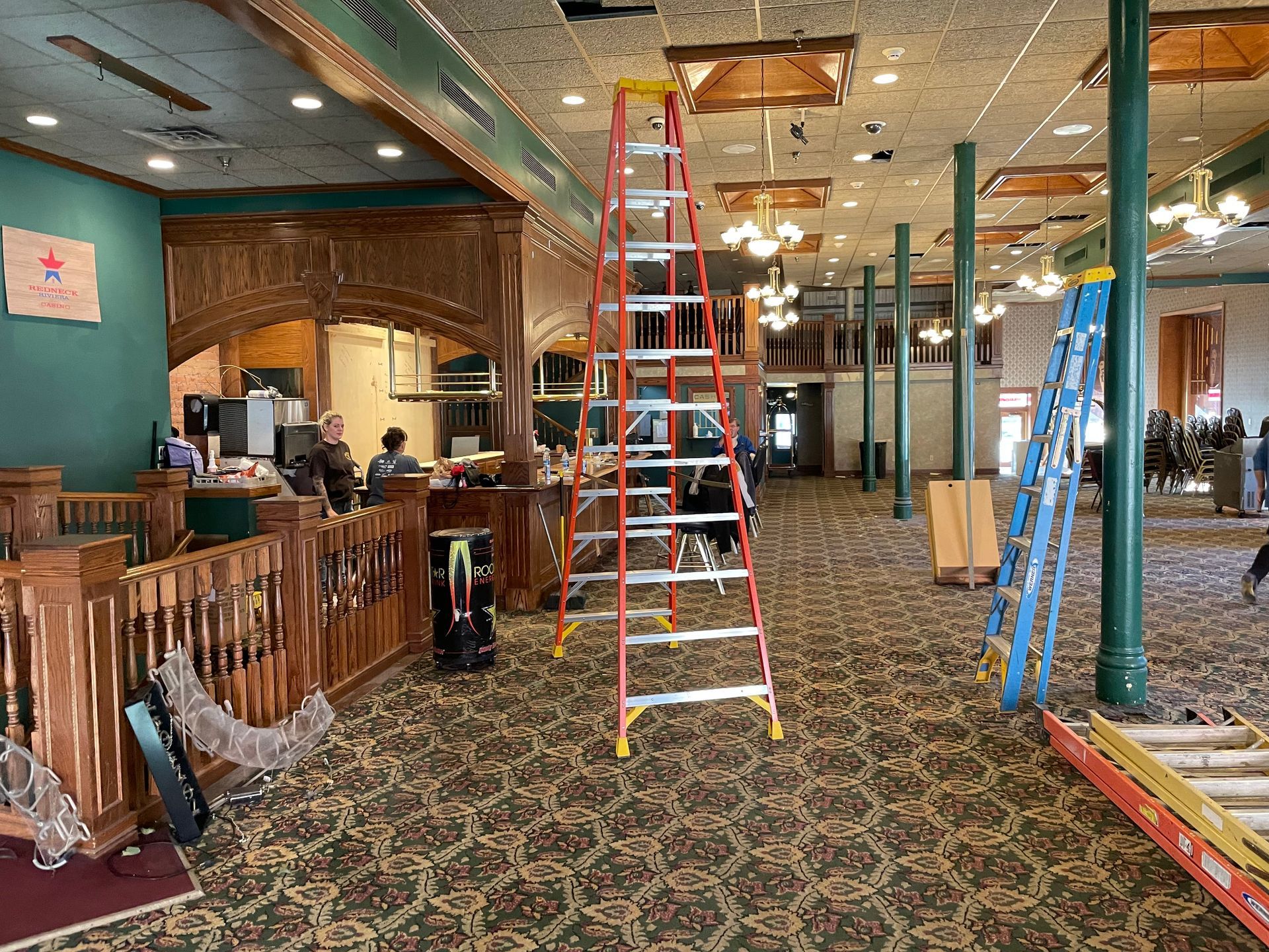 Interior of a hall with ladders, a counter, and workers present, possibly under renovation.