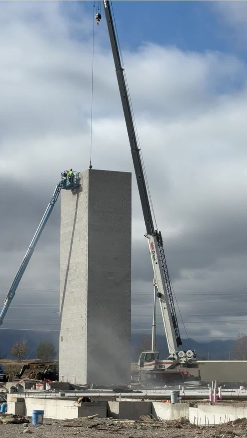 Construction crane lifting a tall, gray concrete pillar. Workers on lift platform, sunny day.