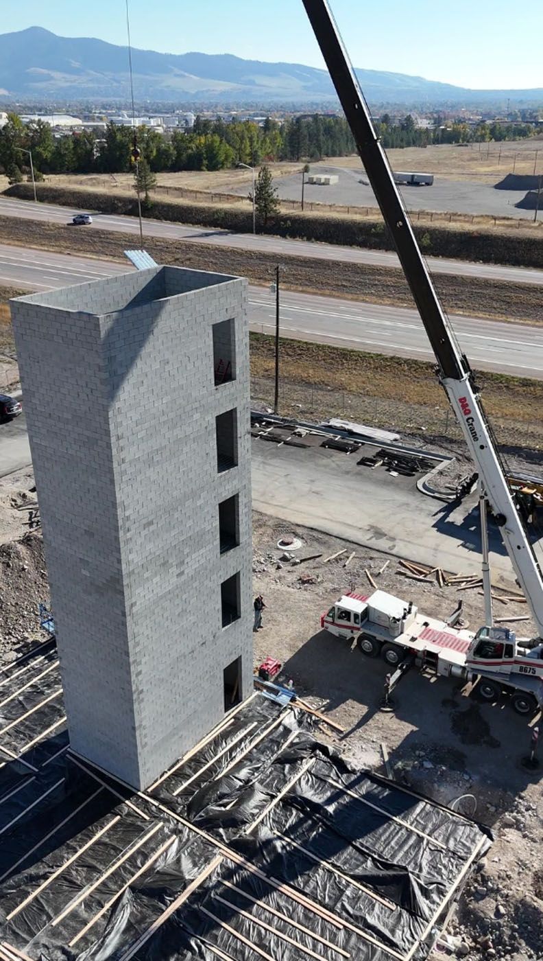 A tall gray concrete structure with window openings is being lifted by a crane at a construction site.