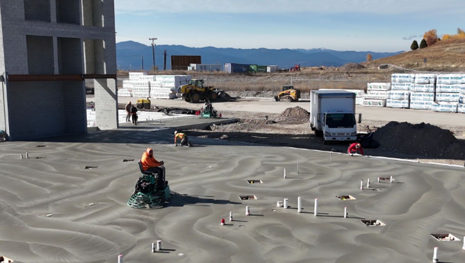 Construction site: worker operating a concrete finishing machine. Building under construction, truck, and equipment visible.