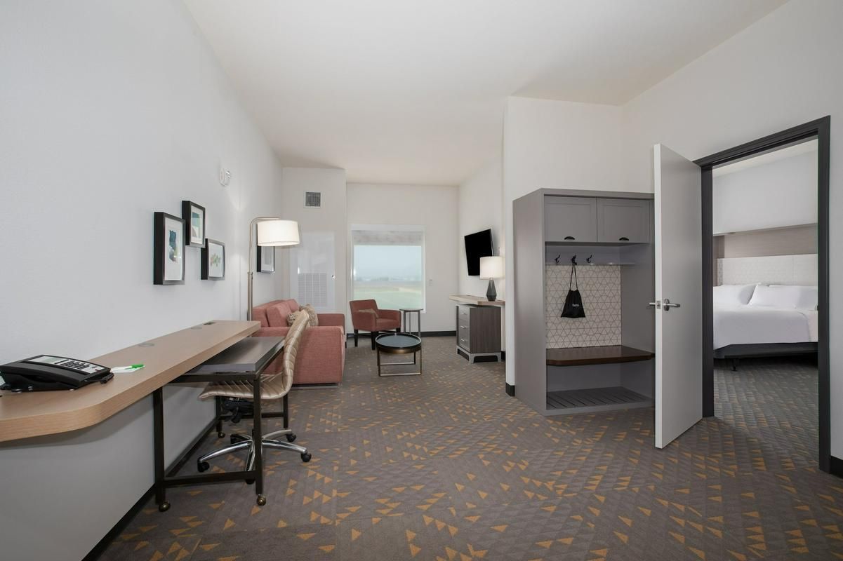 Hotel suite with a living area, desk, and view into the bedroom. White walls, carpet, and a pink chair.
