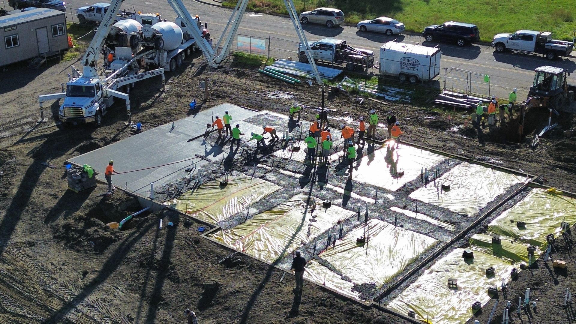 Construction site: Workers pour concrete foundation; trucks, equipment, and vehicles surround the site.