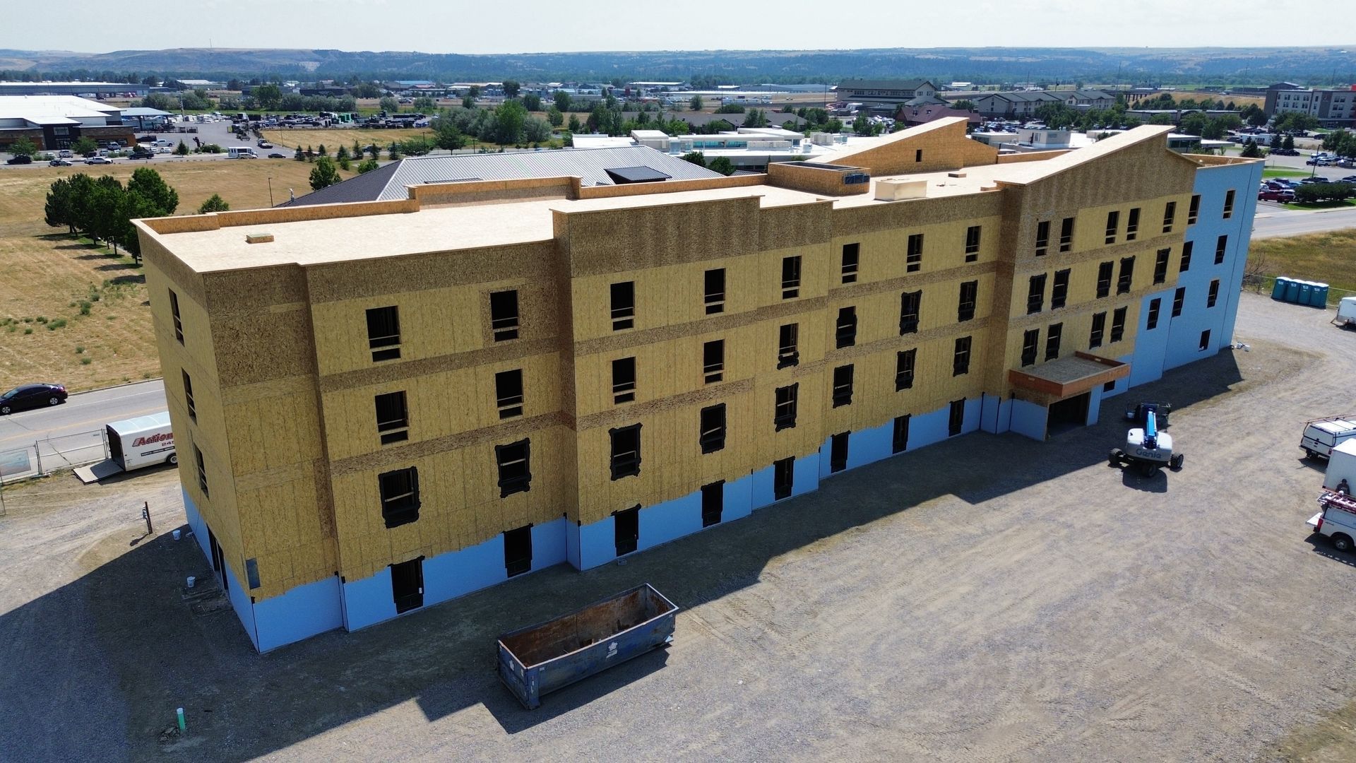 Building under construction, three stories, wood and blue exterior, gravel lot, sunny day.