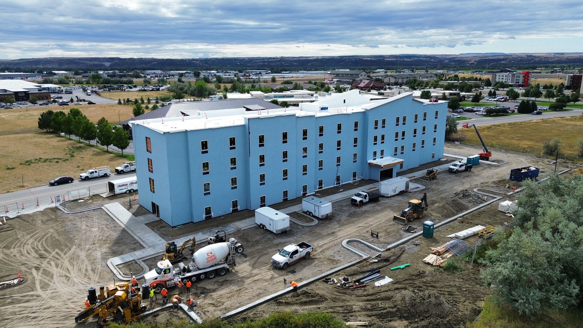 Blue building under construction with construction vehicles and workers on-site, a cloudy sky in the background.