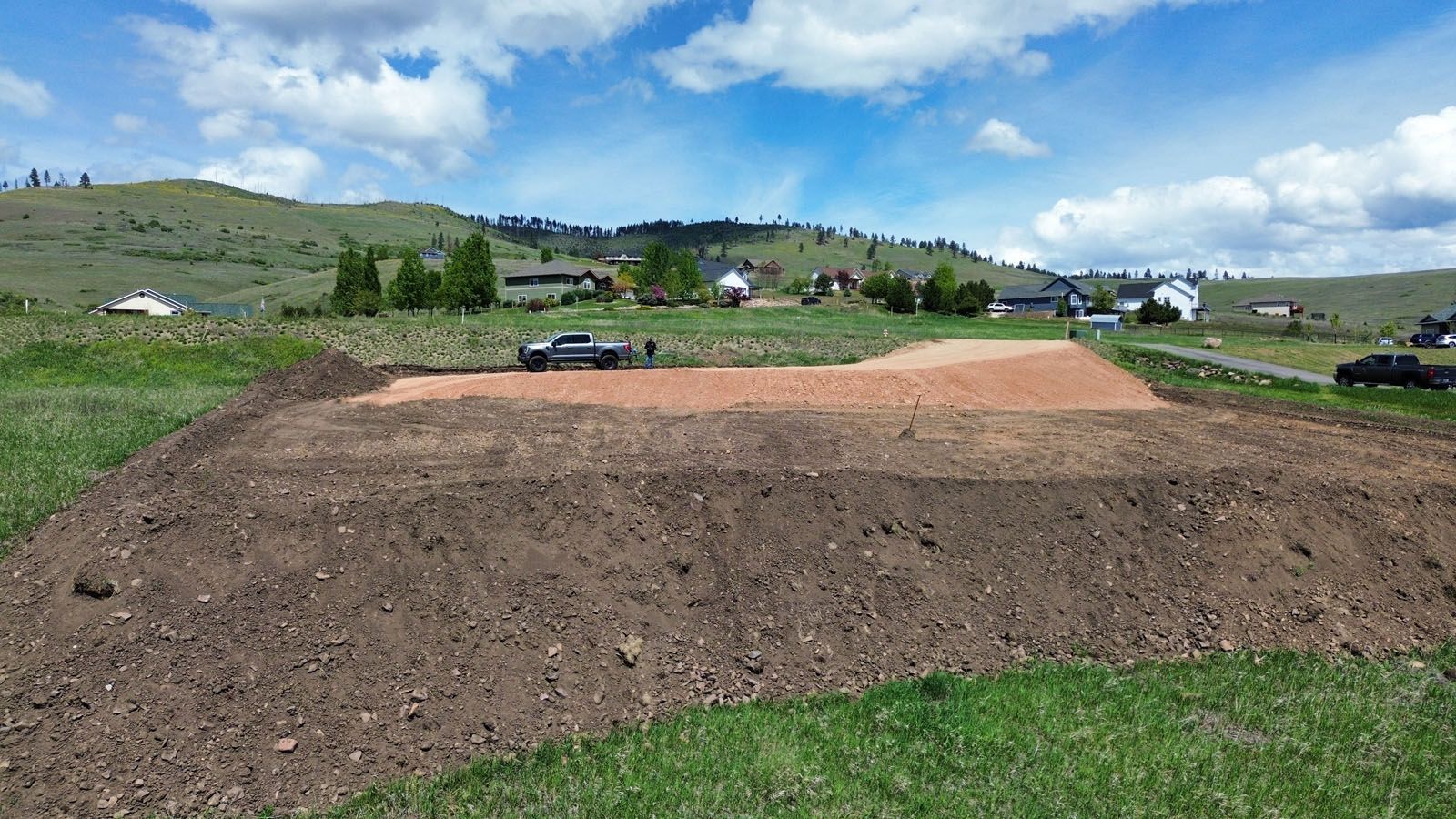 Dirt work with a truck, prepared land, green grass, rolling hills and houses under a partly cloudy sky.