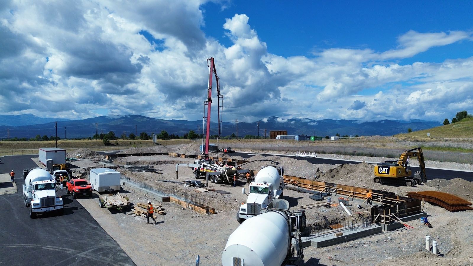 Construction site with cement trucks, pump, and excavator under a cloudy blue sky, mountains in the background.