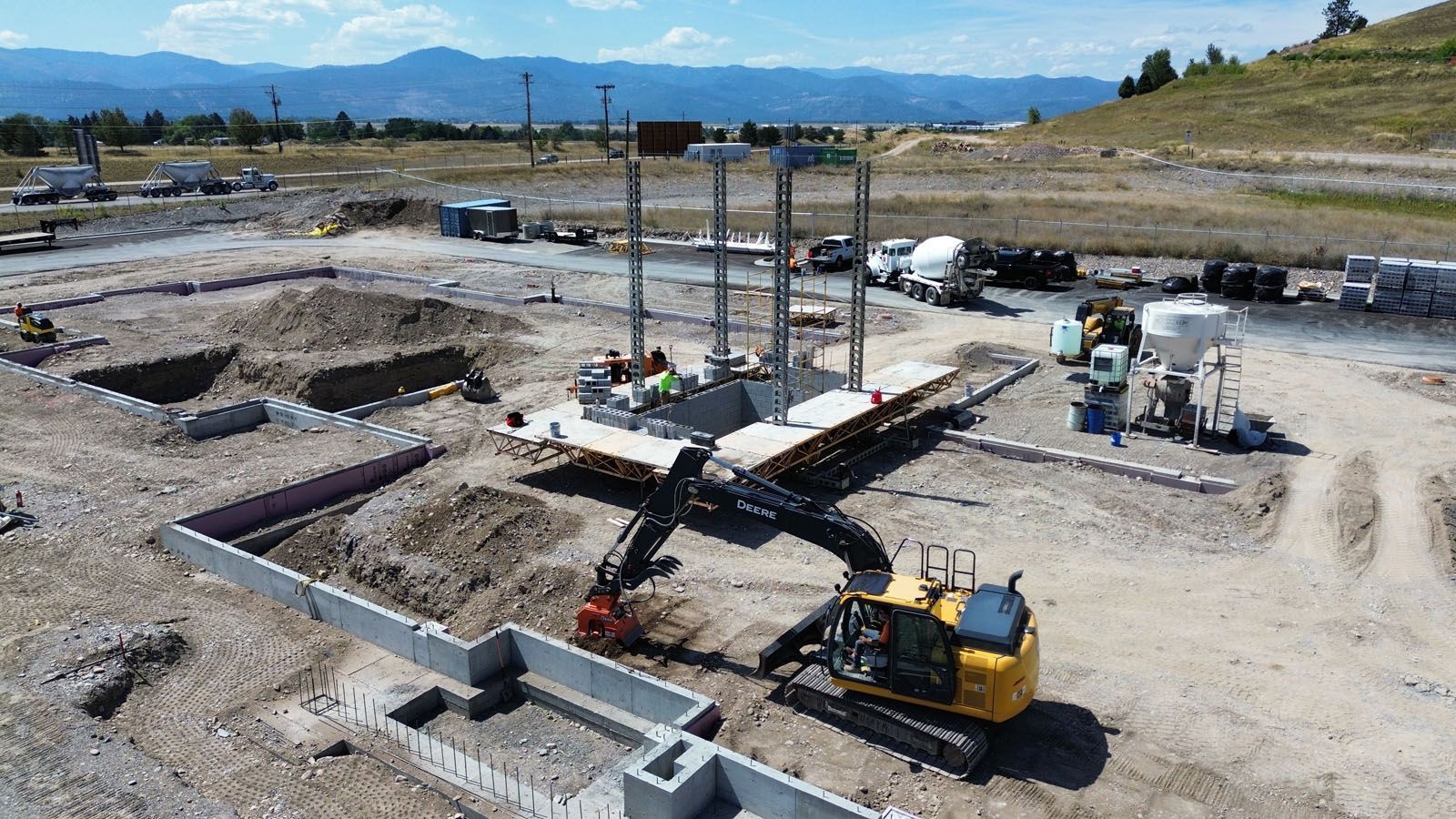 Construction site with excavator and workers around concrete foundations and pillars.