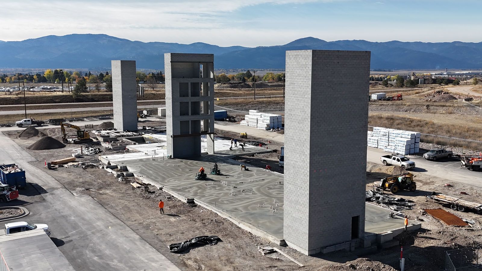 Construction site with three tall concrete structures, mountains in the background, clear skies.