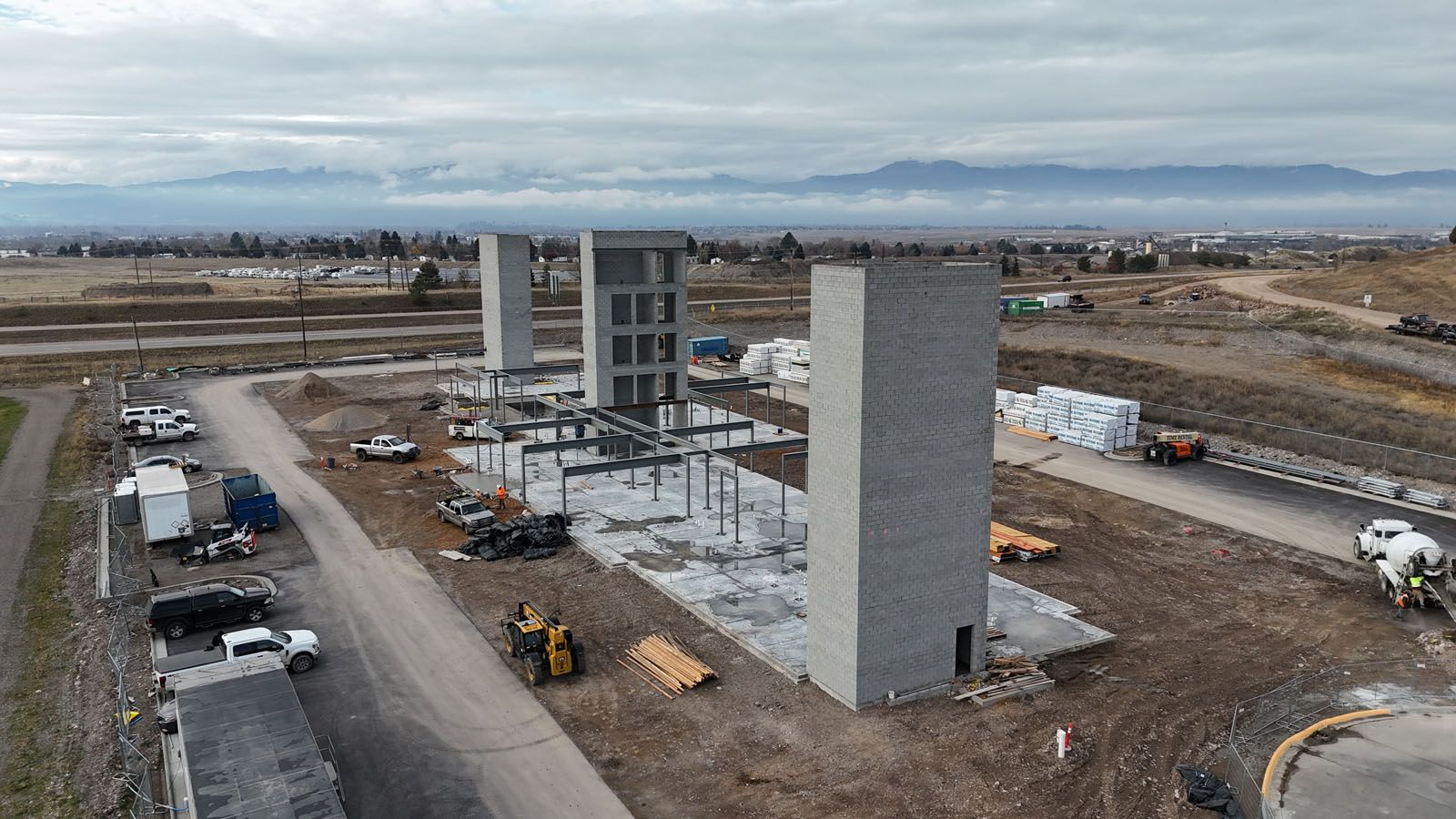Construction site with three large concrete structures, metal framework, and construction vehicles. Overcast sky, distant mountains.