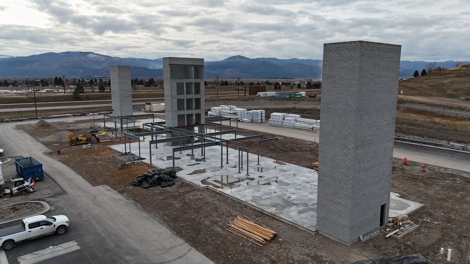 Construction site with three tall concrete towers and steel framework, with mountains in the background.