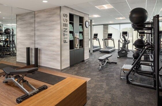 Fitness room with exercise equipment: treadmills, elliptical, weights, and bench, with towels on a shelf.