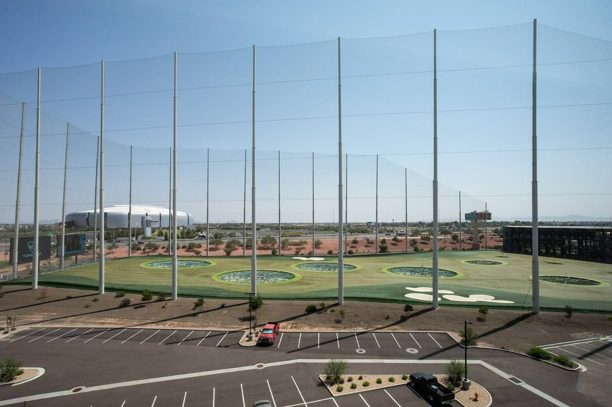 A golf driving range with tall netting and a stadium visible in the distance.