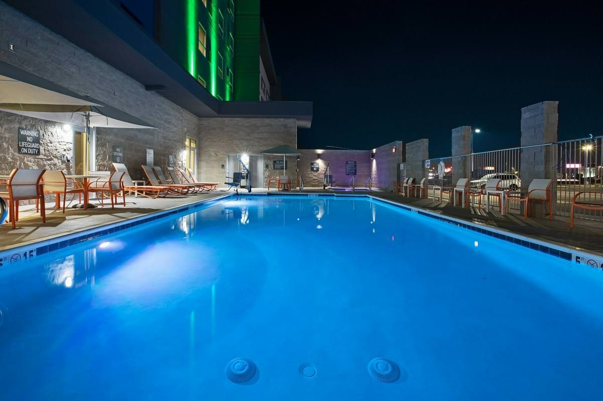 Rooftop pool at night, lit with blue lights. Lounge chairs and a building with green lights are nearby.
