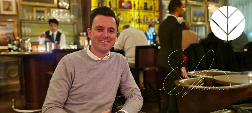 Man smiles in a restaurant; bar and staff in background. A logo is on the top right.