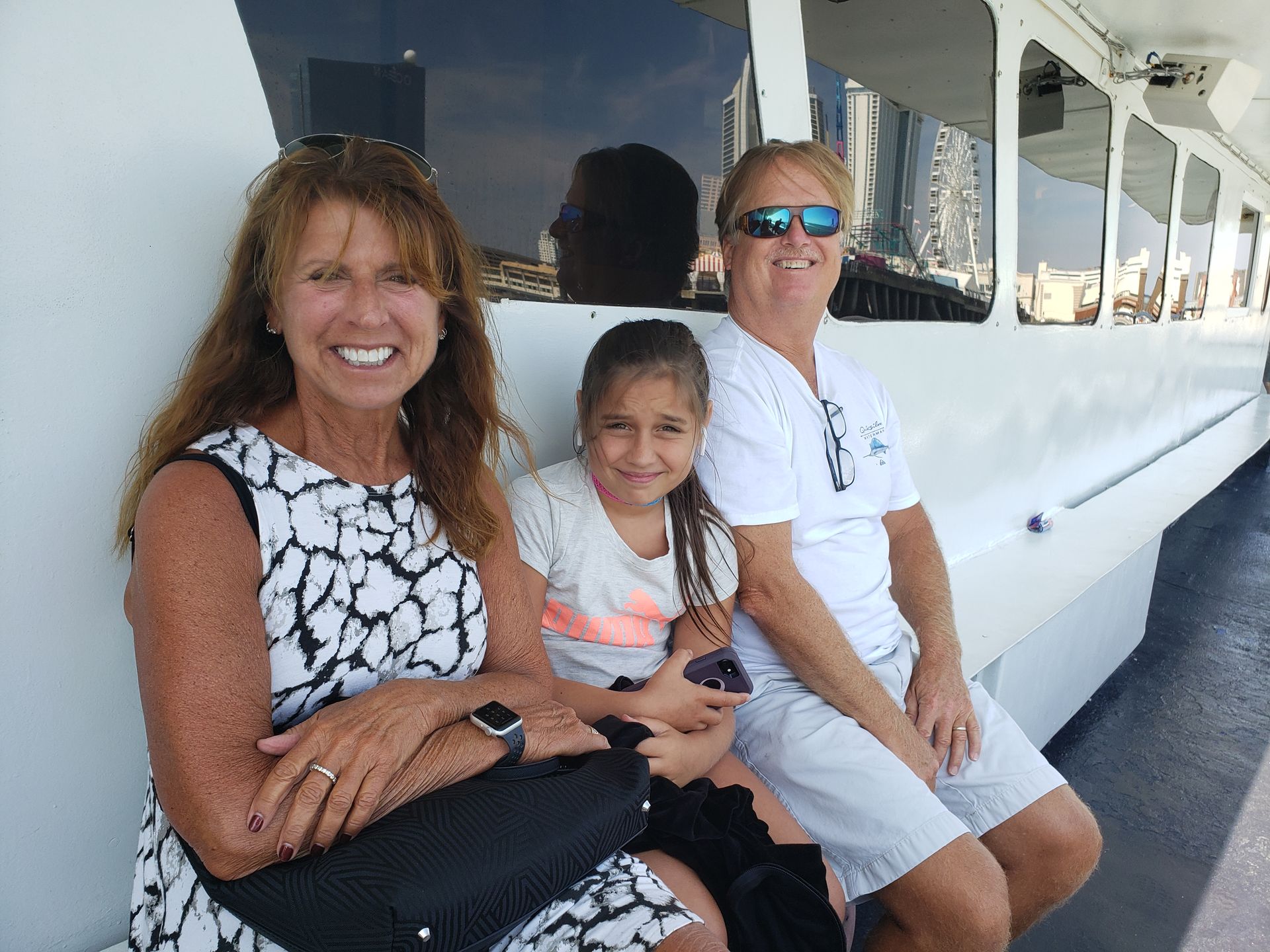 Three people smiling on a boat: woman in patterned dress, girl with phone, and person in sunglasses.