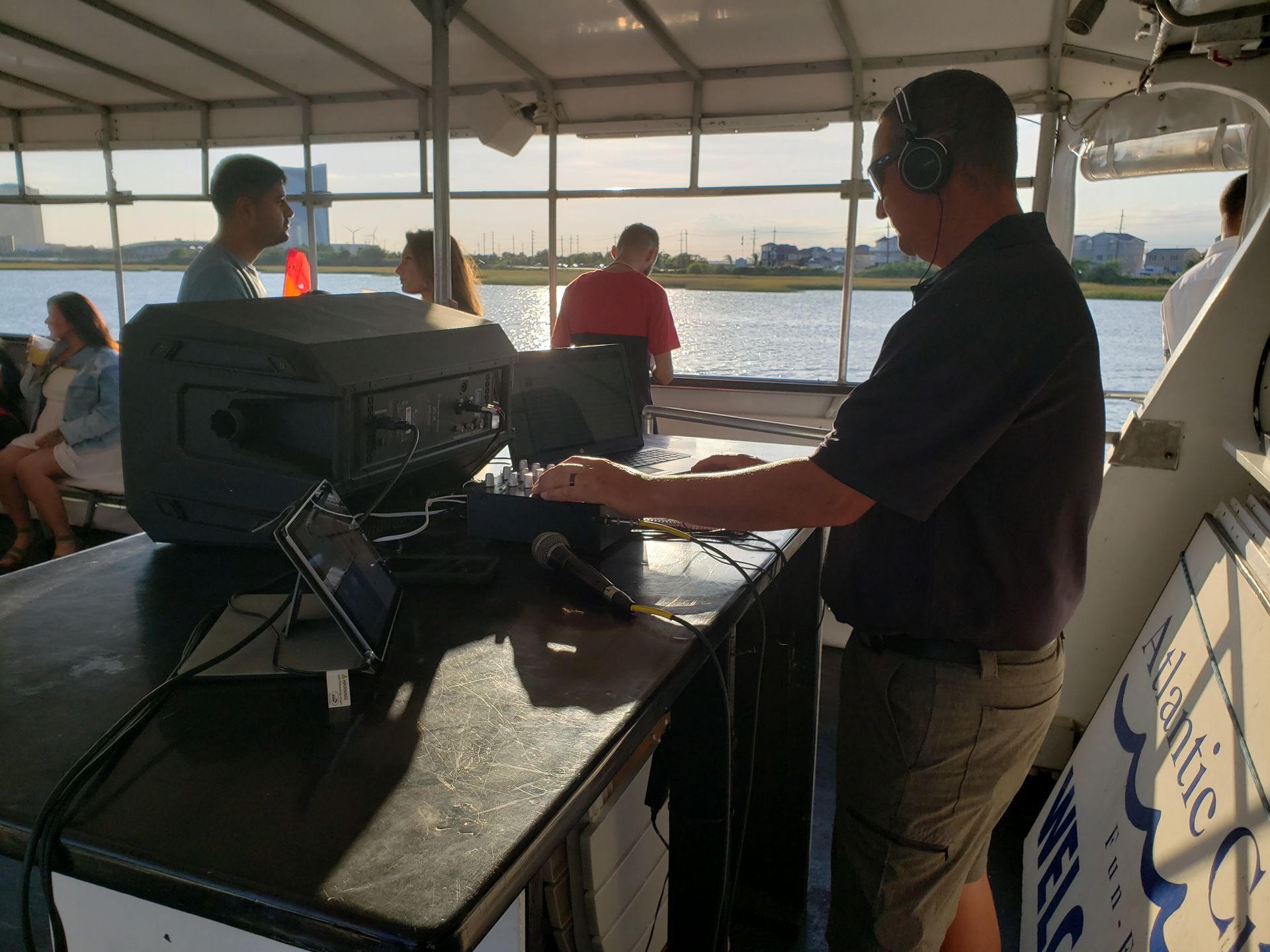 Man operating DJ equipment on a boat, overlooking water and buildings, with people in background.
