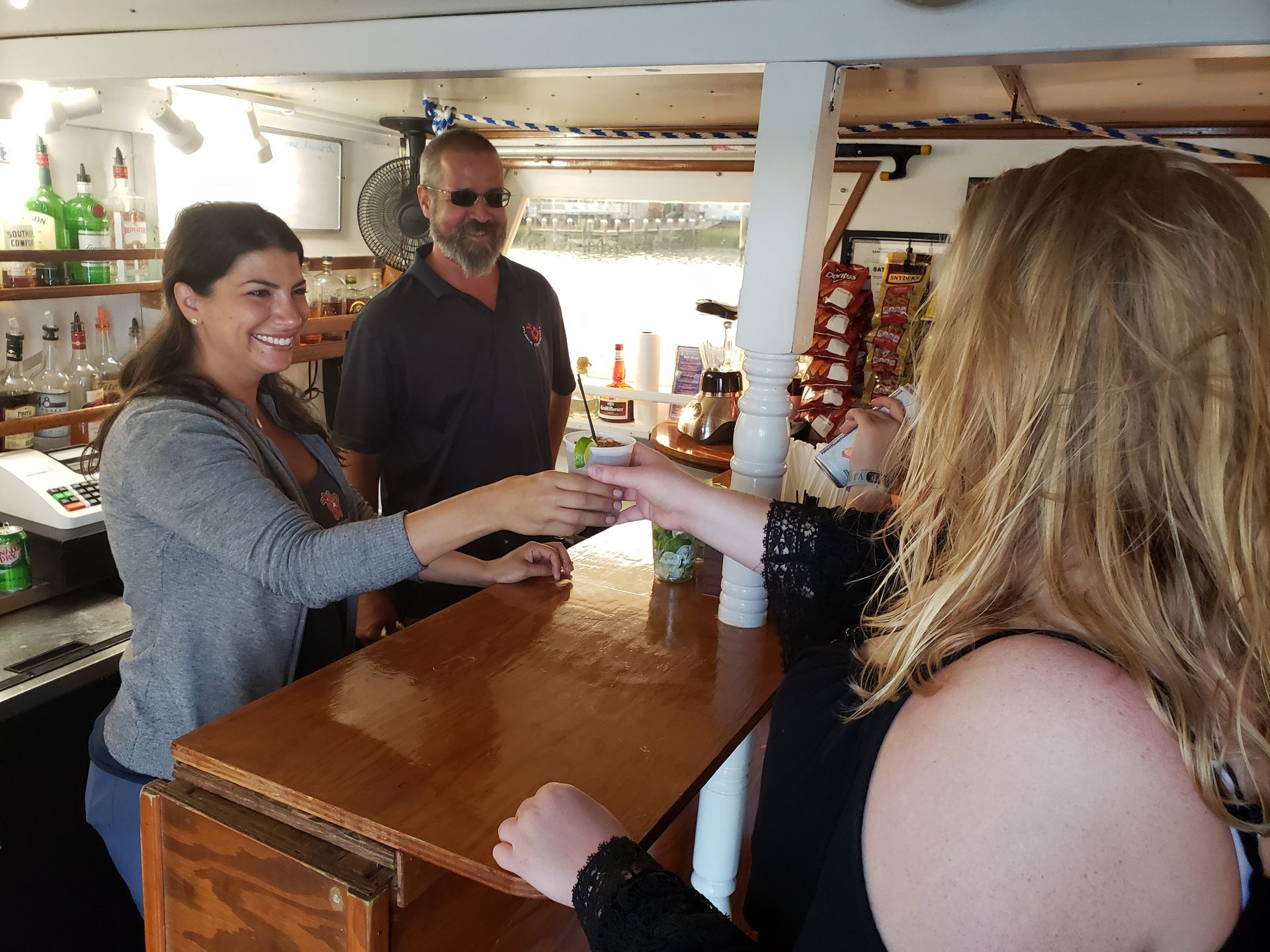 Woman handing drink to another woman at a bar, man in background. Bar is inside with shelves of liquor.