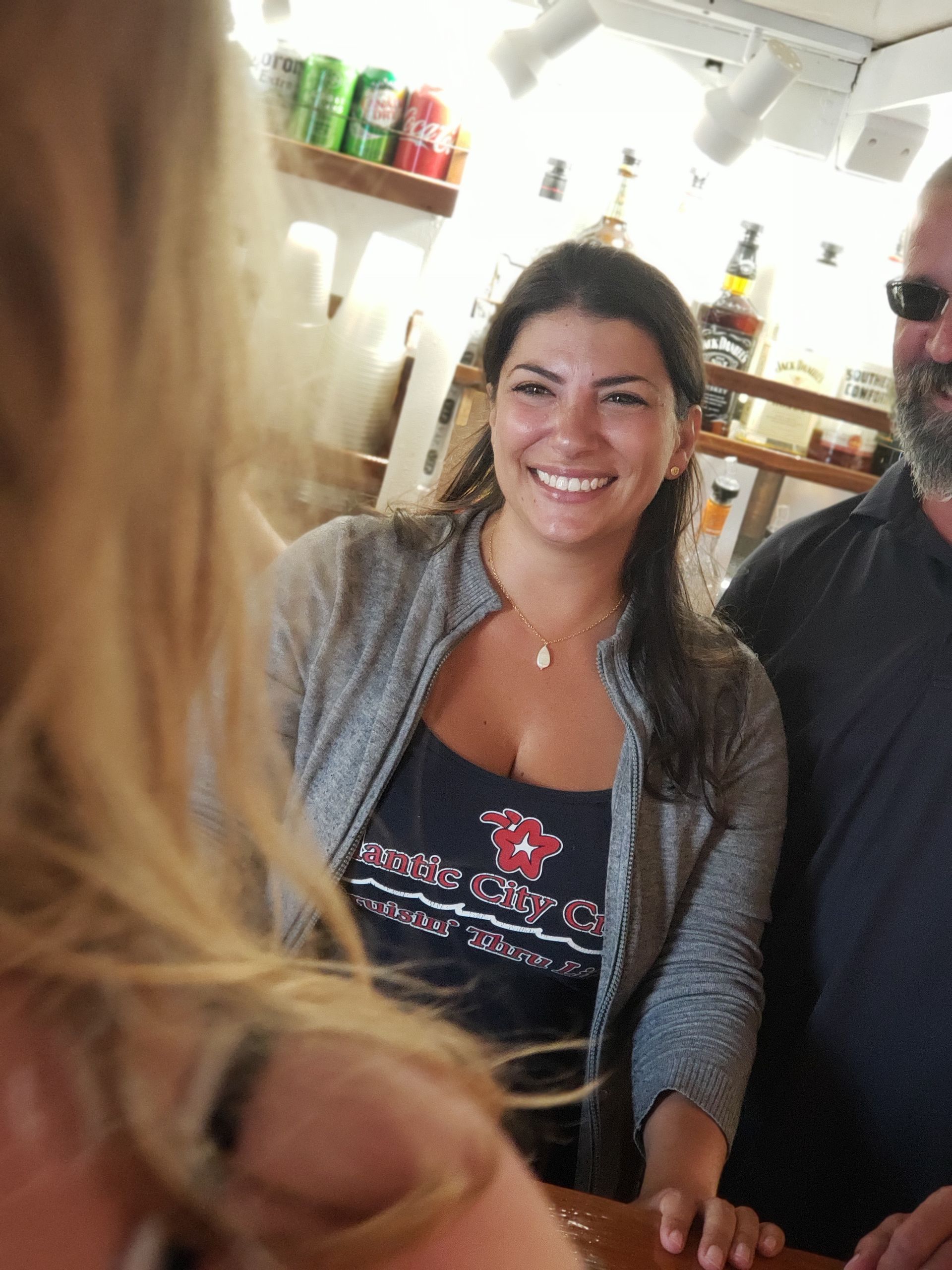 Woman smiling, wearing a gray sweater over a tank top at a bar.