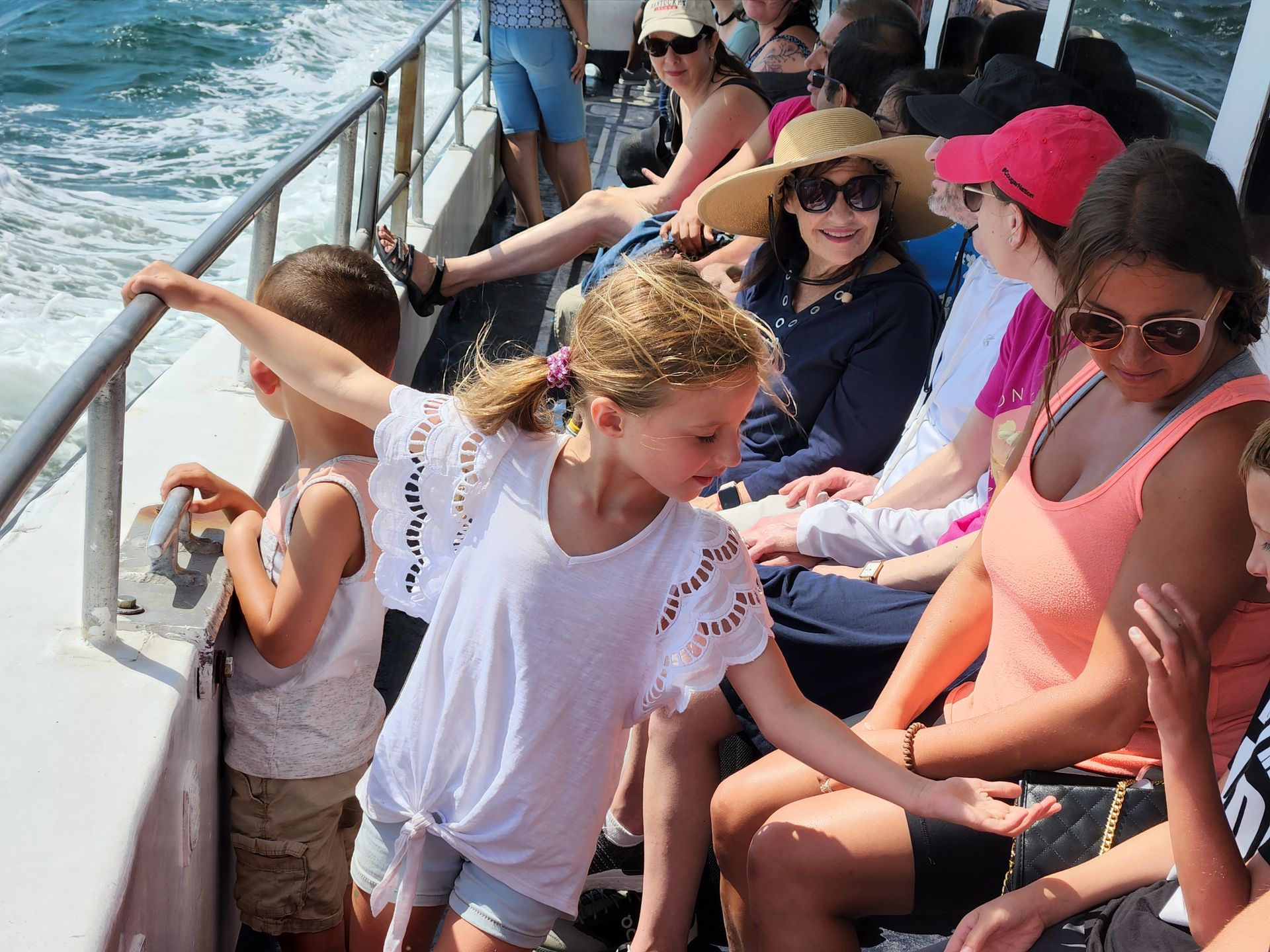 People on a boat looking at the water. A girl in a white shirt reaches out. Sunny day.