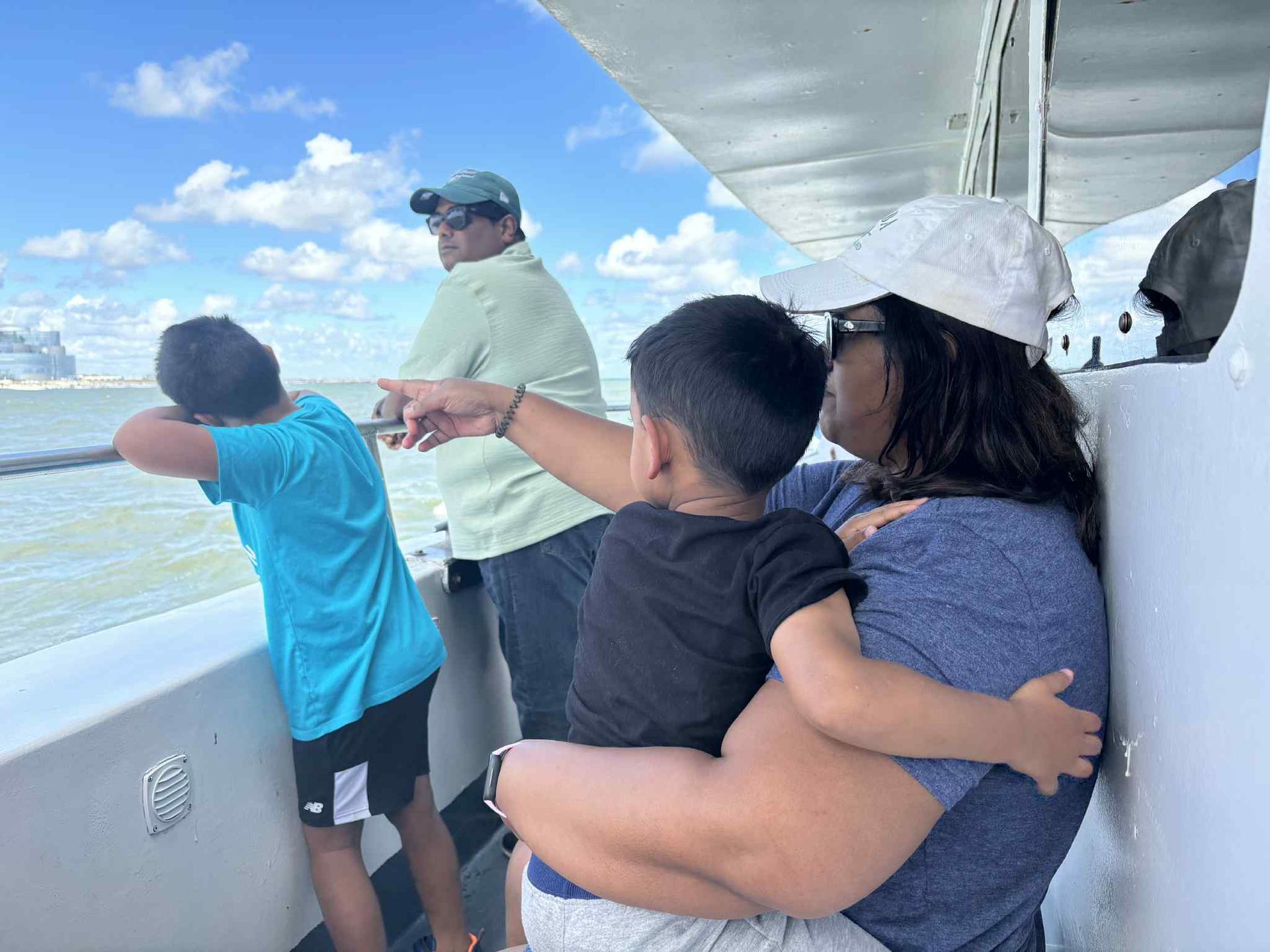 Family on a boat, looking out at the water. Child points, others gaze at horizon under a cloudy sky.