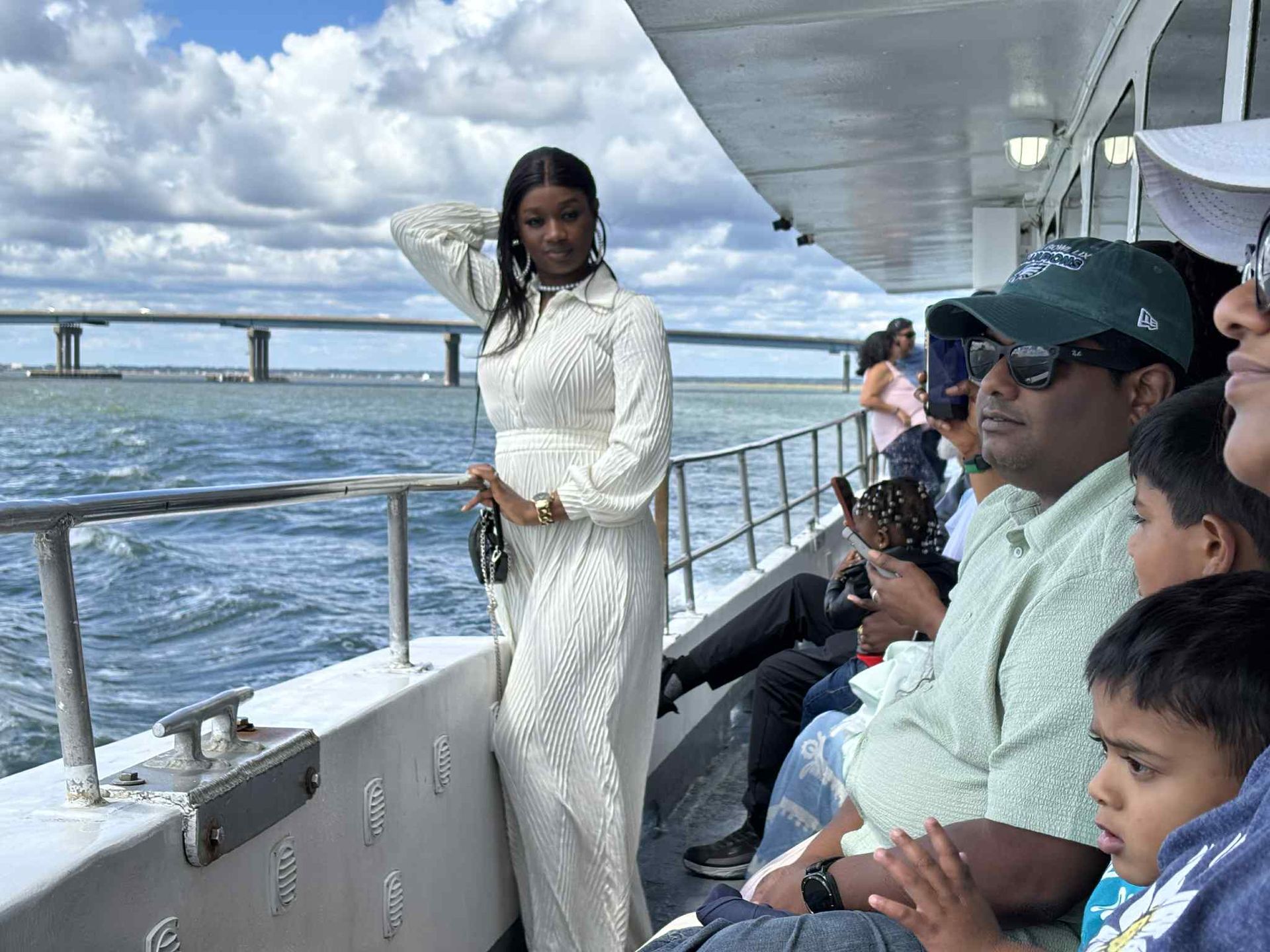 Woman in white dress on boat, looking at water, bridge in background, people seated nearby.