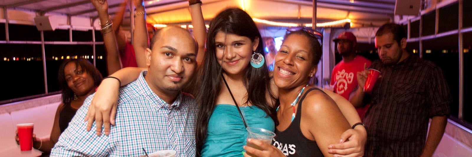 Group of friends smiling and embracing on a boat at night. They are holding drinks, and some are raising their arms.