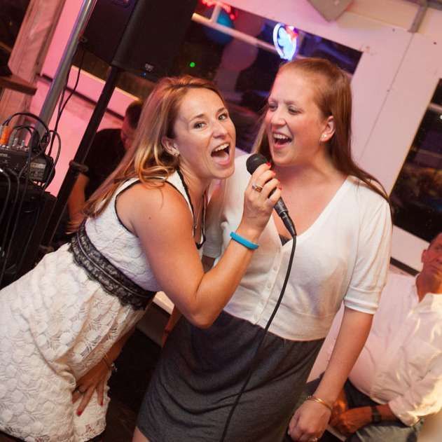 Two women singing into a microphone at a karaoke bar; one in a white dress, the other in a white top and grey skirt.