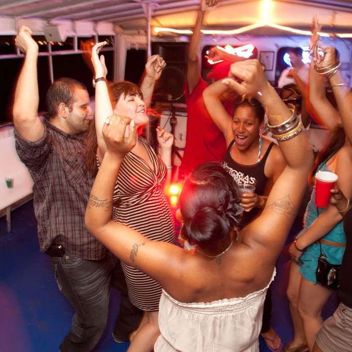 People dancing on a boat, arms raised. Party atmosphere with bright lights.