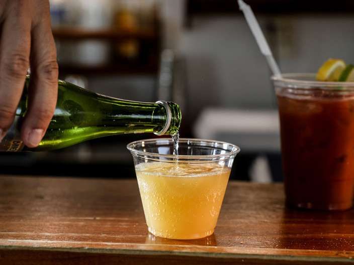 Bartender pours champagne into a plastic cup; a bloody mary sits beside it.