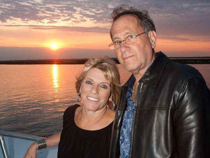 Couple on a boat at sunset; woman smiles, man wears glasses, leather jacket.