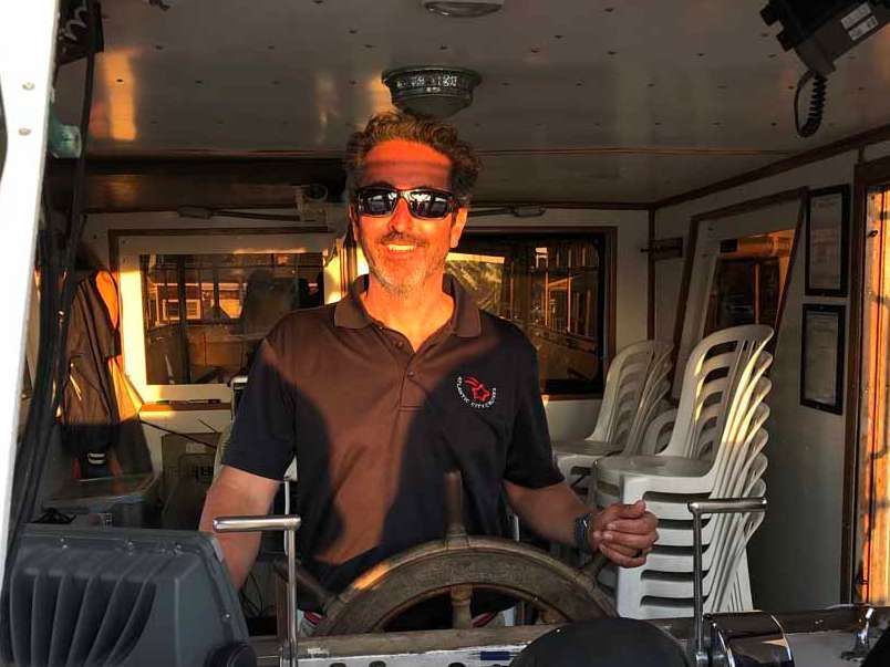 Man wearing sunglasses at the helm of a boat, with stacked white chairs visible behind him.