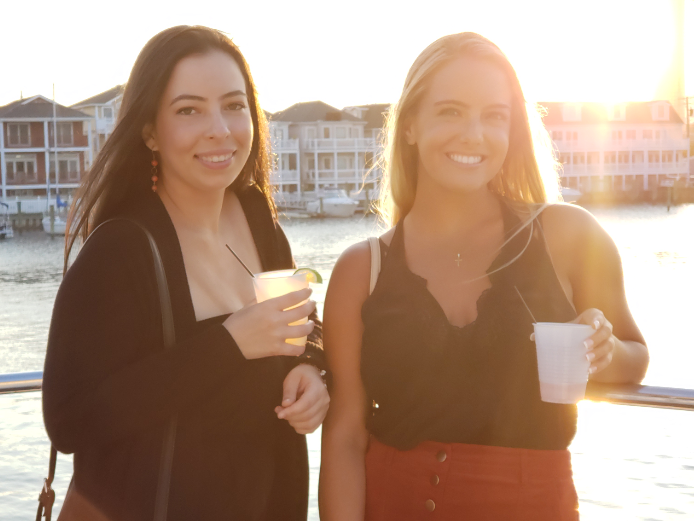 Two women smile, holding drinks, on a boat at sunset.