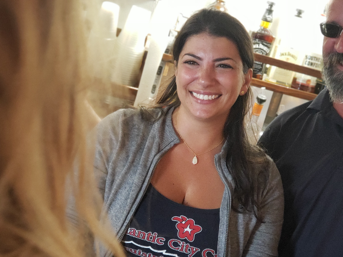 Woman smiling, wearing a gray cardigan over a dark shirt with a logo, standing near a man with a beard.