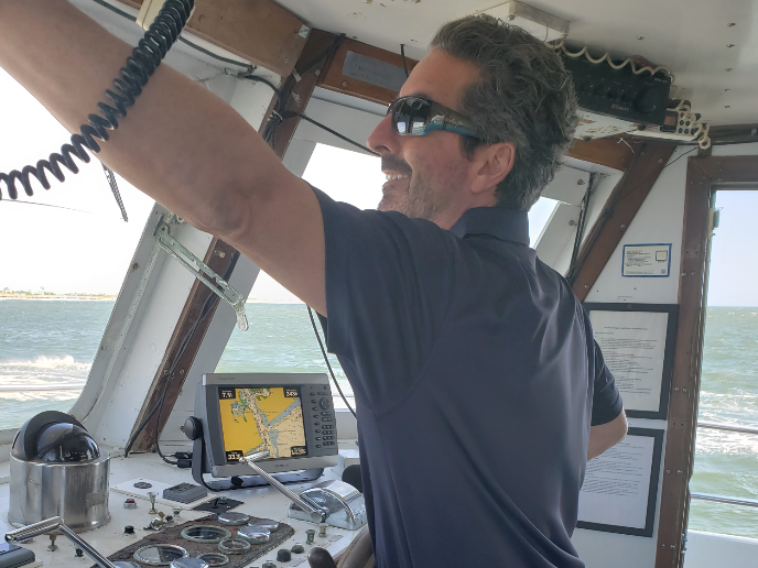 Man in sunglasses steers a boat. Inside a cabin, arm raised, looking ahead.