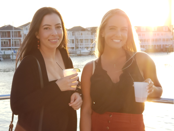 Two women smiling, holding drinks, in front of a waterfront setting at sunset.