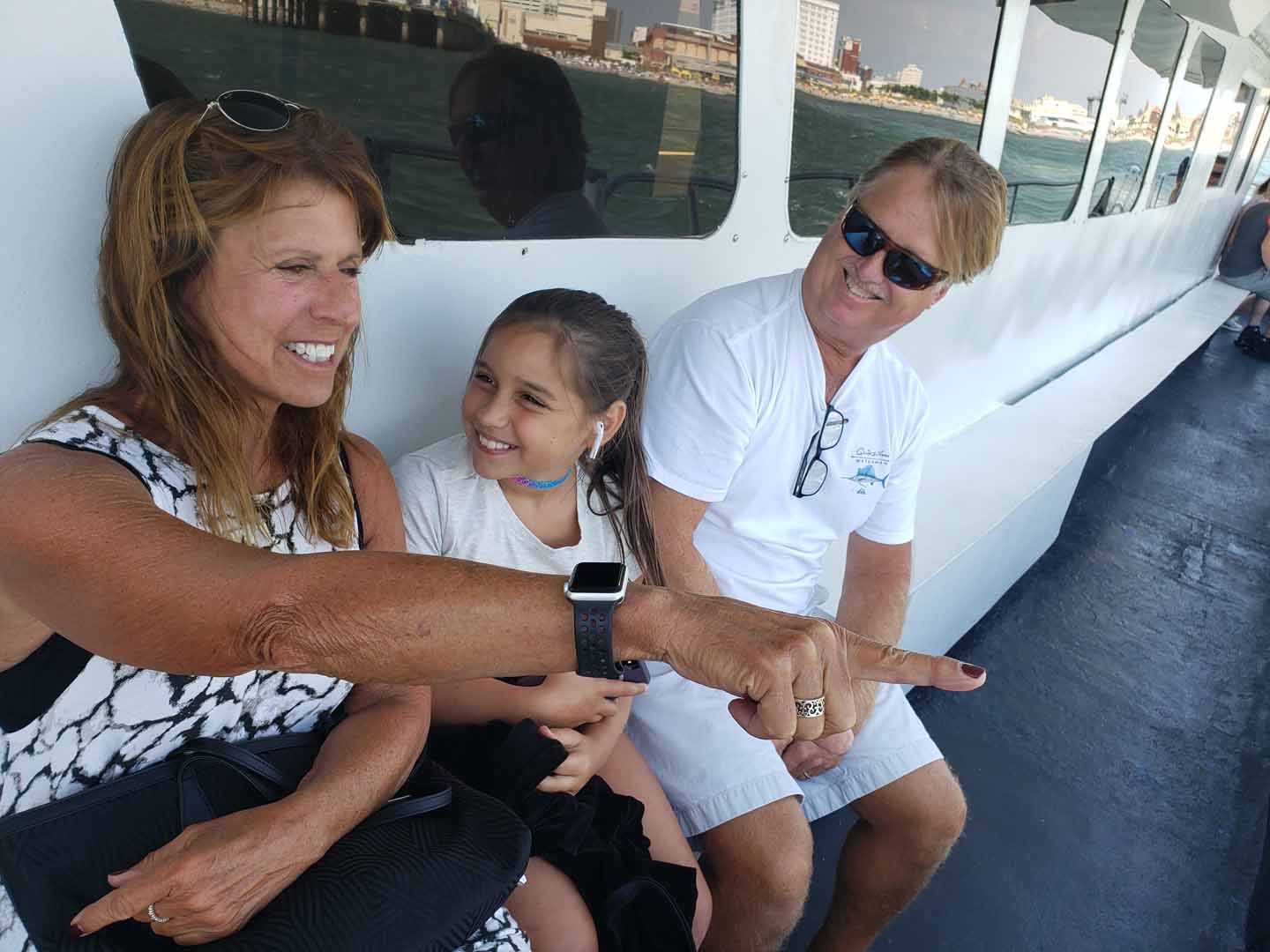 Woman points, smiling, to something out of frame. Girl and man smile on boat. Blue water and cityscape visible.