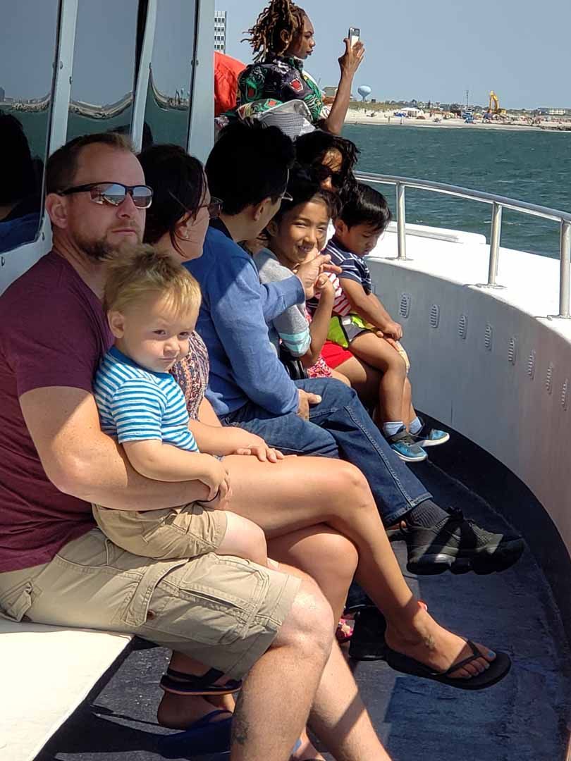 People on boat, looking at the water. Man holding child, others seated. Sunny day.