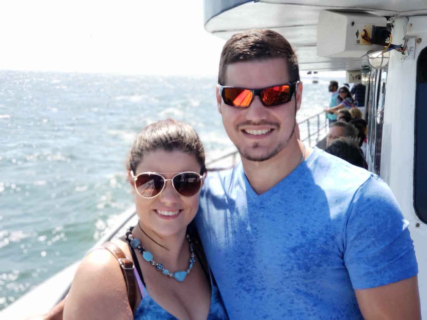 Couple on boat, smiling at camera. Blue water and sky background. Man in red sunglasses, woman in tan sunglasses.
