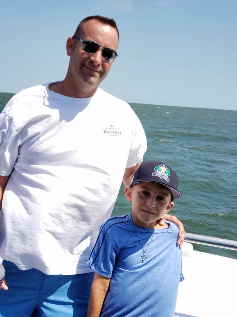 Man and boy on a boat, posing by ocean. Man wearing sunglasses and white shirt, boy wearing baseball cap and blue shirt.