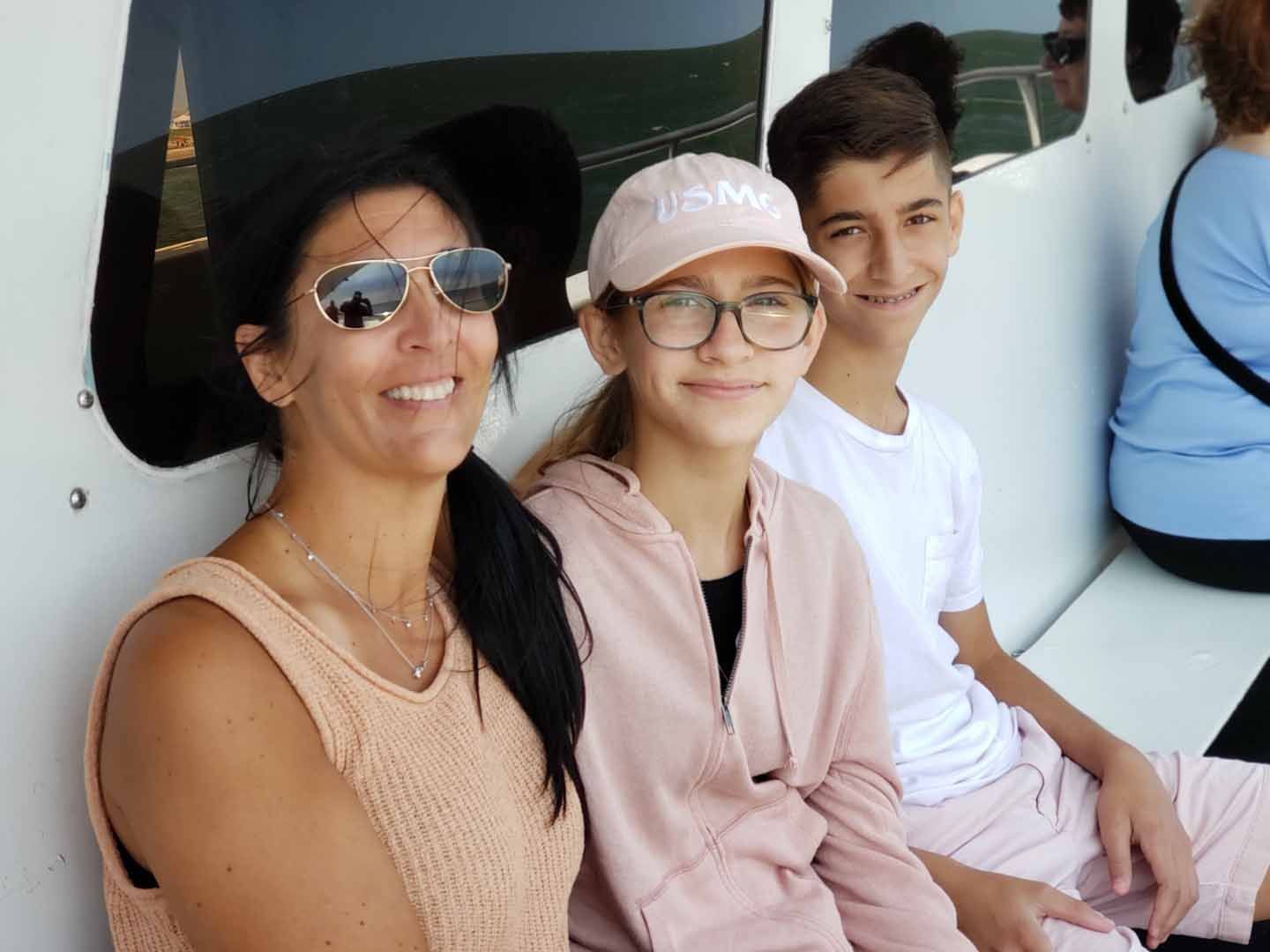 Woman and two teens smiling, sitting inside a vehicle with a white interior; sunny day.
