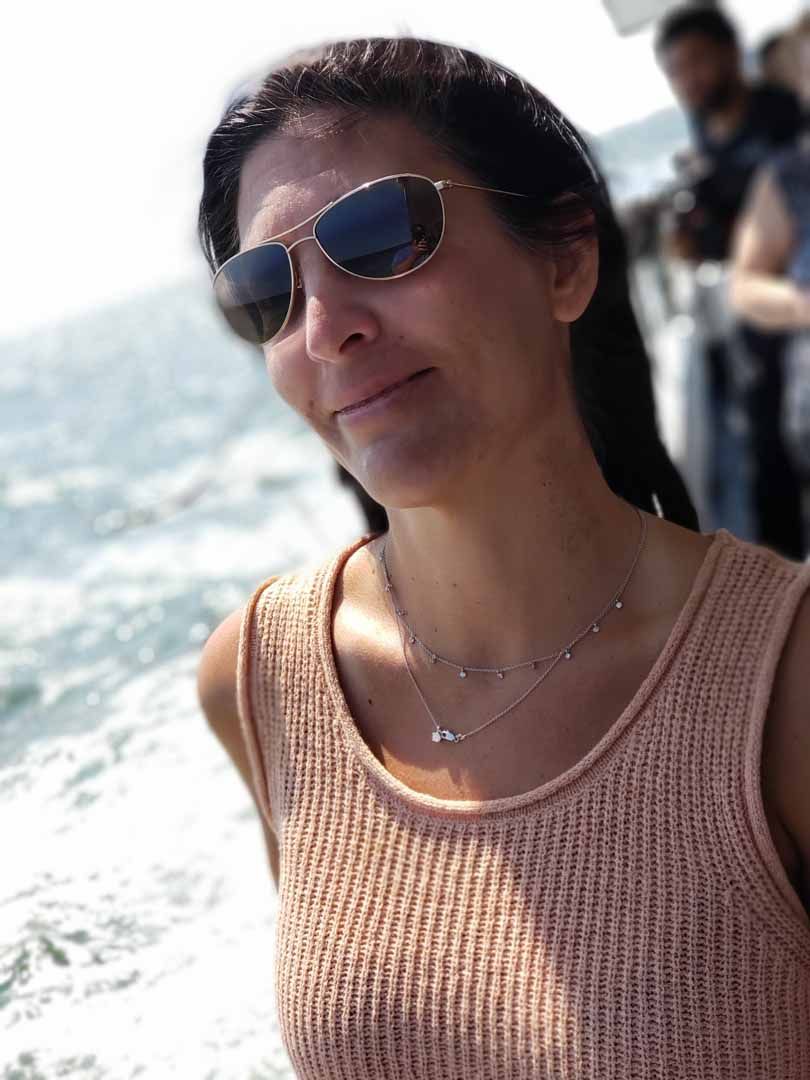 Woman wearing sunglasses on a boat, smiling, with water and people in the background.