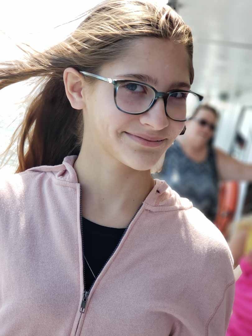 Girl wearing glasses and a pink hoodie smiles on a boat, hair blowing in the wind.