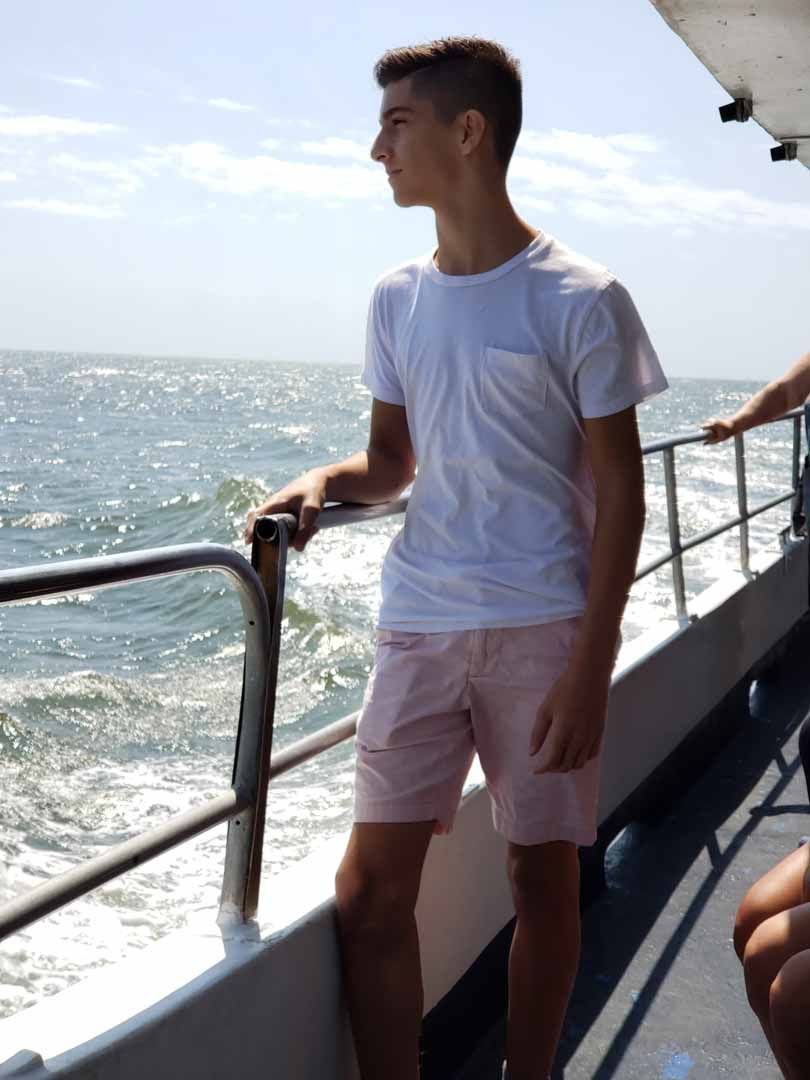 Man on a boat, looking at the ocean. He wears a white shirt and pink shorts.