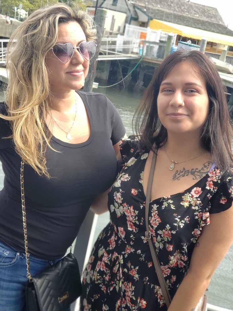 Two women near a pier, one with sunglasses, the other in a floral dress, both smiling.