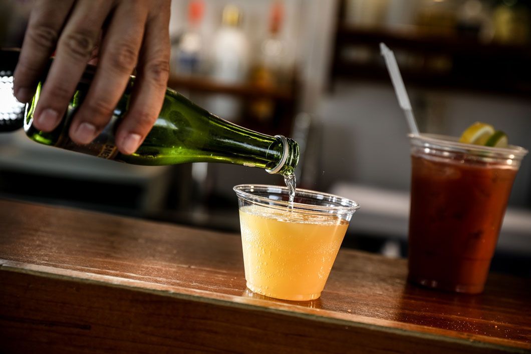 Bartender pours champagne into a clear plastic cup on a bar, next to a red Bloody Mary.