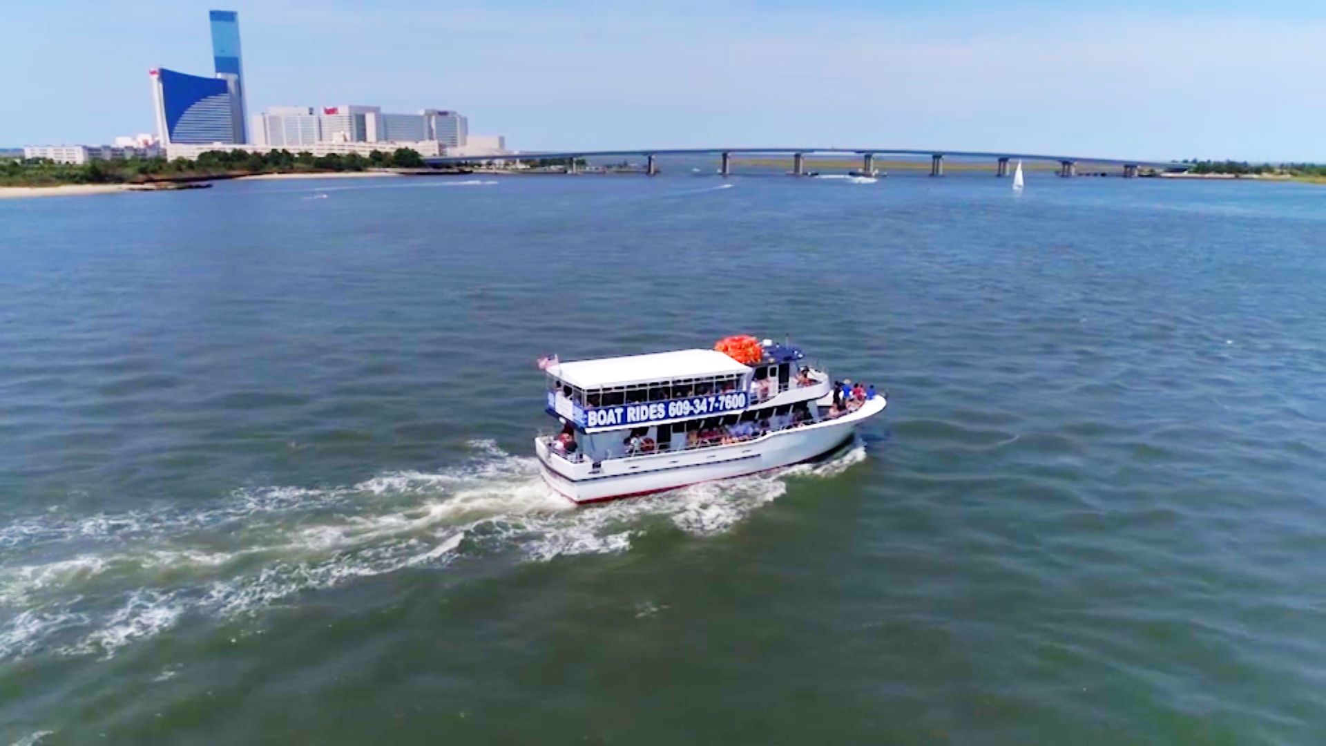 A boat with passengers on water, buildings and a bridge in the background, blue sky.