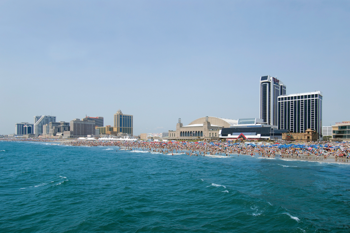 Beach filled with people; buildings in the distance. Blue ocean, clear sky.